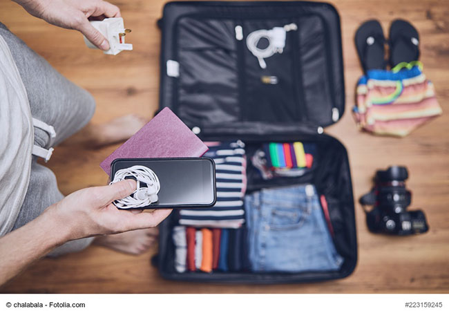 Junger Mann packt Koffer und überlegt, was darf ins Handgepäck – das Ladegerät, das Smartphone? Foto: Fotolia © chalabala